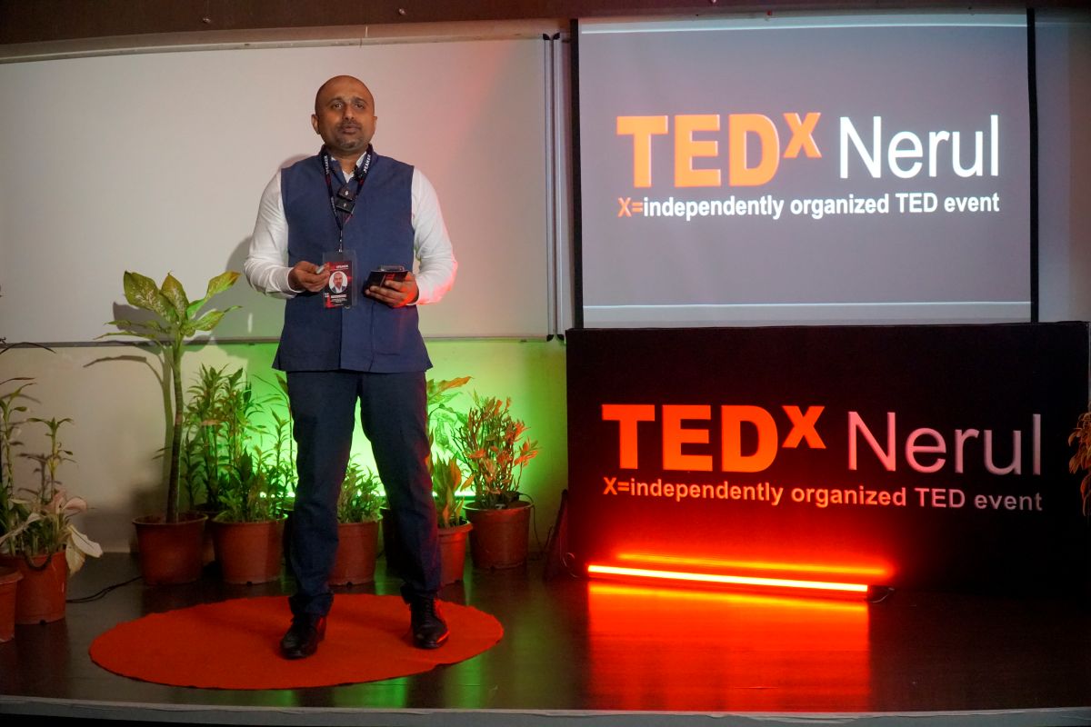 Dr. Mohana Rao Patibandla standing on a red circular carpet on stage at the TEDx Nerul event, wearing a blue vest and white shirt, holding a microphone. The backdrop features the TEDx Nerul logo and the text "X=independently organized TED event" on a screen and a neon sign, with green plants in pots around the stage.